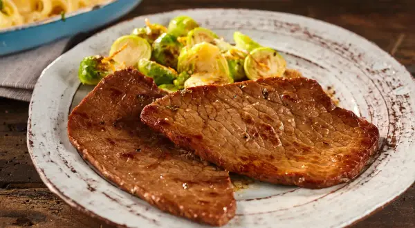 Due fette di paillard di vitello succulento servite con cavolini di Bruxelles arrostiti e un contorno di pasta in un piatto rustico.