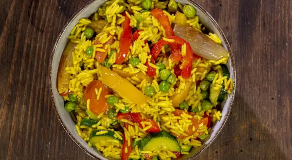 Un invitante riso al curry con piselli, peperoni rossi e gialli, zucchine e cipolla, servito in una ciotola su un tavolo di legno.