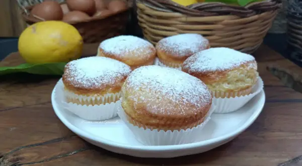Cinque muffin allo yogurt spolverati di zucchero a velo, pronti per essere gustati. Sullo sfondo, limoni e uova fresche.