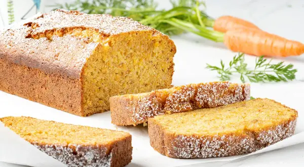 Torta carote e mandorle soffice e dorata, cosparsa di zucchero a velo. Due fette sono servite accanto al pane intero, con carote fresche sullo sfondo.