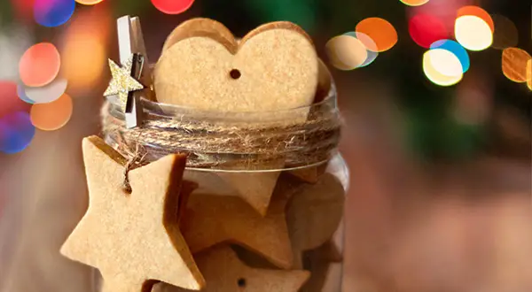 Biscotti alla cannella a forma di stella e cuore in un barattolo, decorati con luci natalizie sfocate.