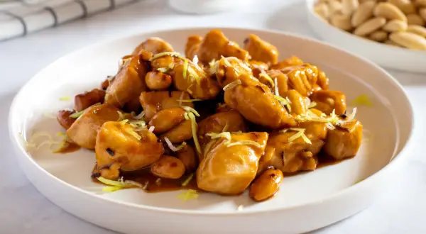 Un invitante piatto di pollo alle mandorle, lucido e dorato, guarnito con erba cipollina. Sullo sfondo, una ciotola di mandorle intere.