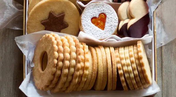 Scatola piena di deliziosi dolci senza glutine: biscotti rotondi con stella al cioccolato, a forma di cuore con zucchero a velo, ricoperti di cioccolato e impilati in file ordinate.