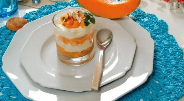 Delizioso semifreddo al melone a strati, guarnito con frutta fresca e mandorle, servito in un bicchiere su un piatto bianco.