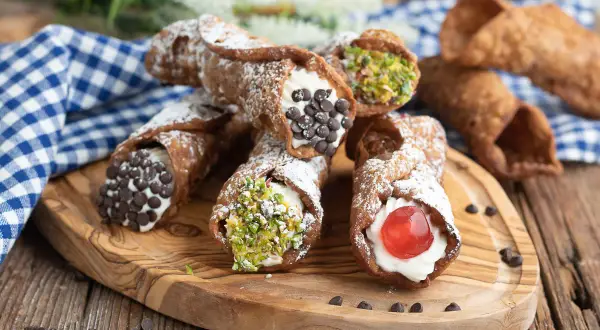 Cannoli siciliani ricetta: deliziosi dolci con crema, gocce di cioccolato, pistacchi e ciliegina candita su tagliere di legno.