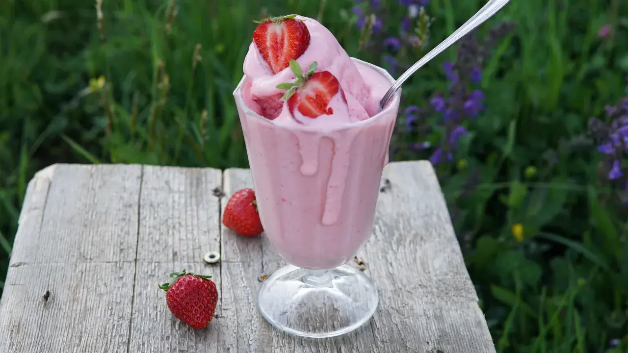 Un cremoso gelato alla fragola, guarnito con fragole fresche, su un tavolo di legno rustico, con un prato fiorito sullo sfondo.