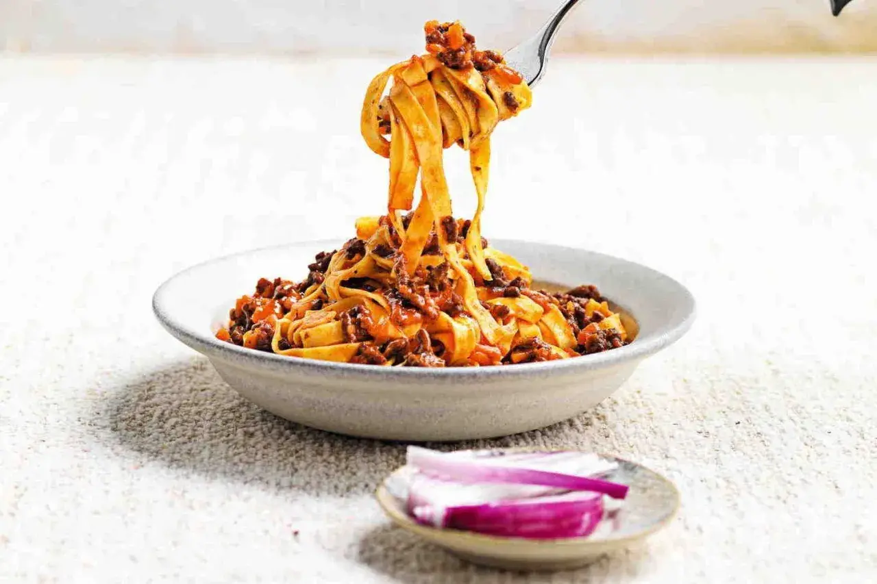 Un piatto invitante di tagliatelle fatte in casa con ragù ricco, sollevate da una forchetta. Accanto, spicchi di cipolla rossa fresca.