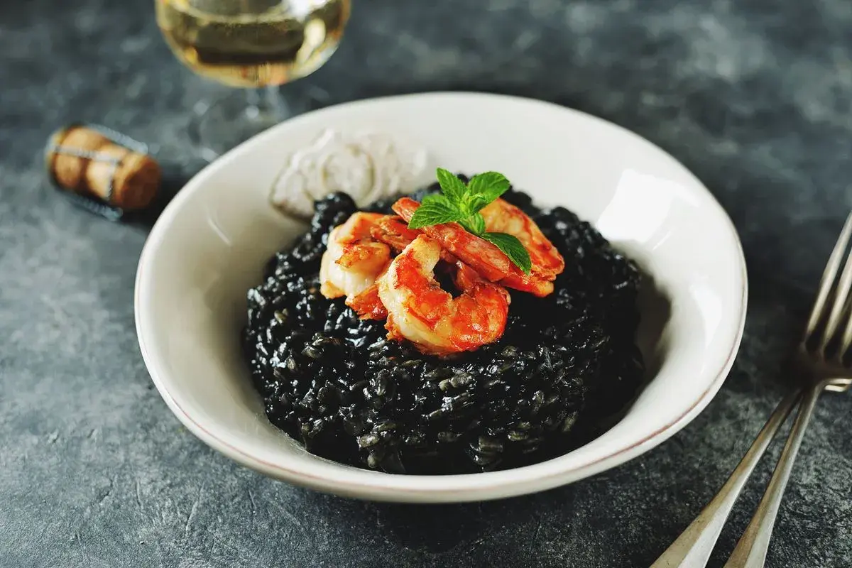 Un invitante risotto al nero di seppia, guarnito con gamberi succulenti e menta fresca, pronto per essere gustato con un bicchiere di vino bianco.