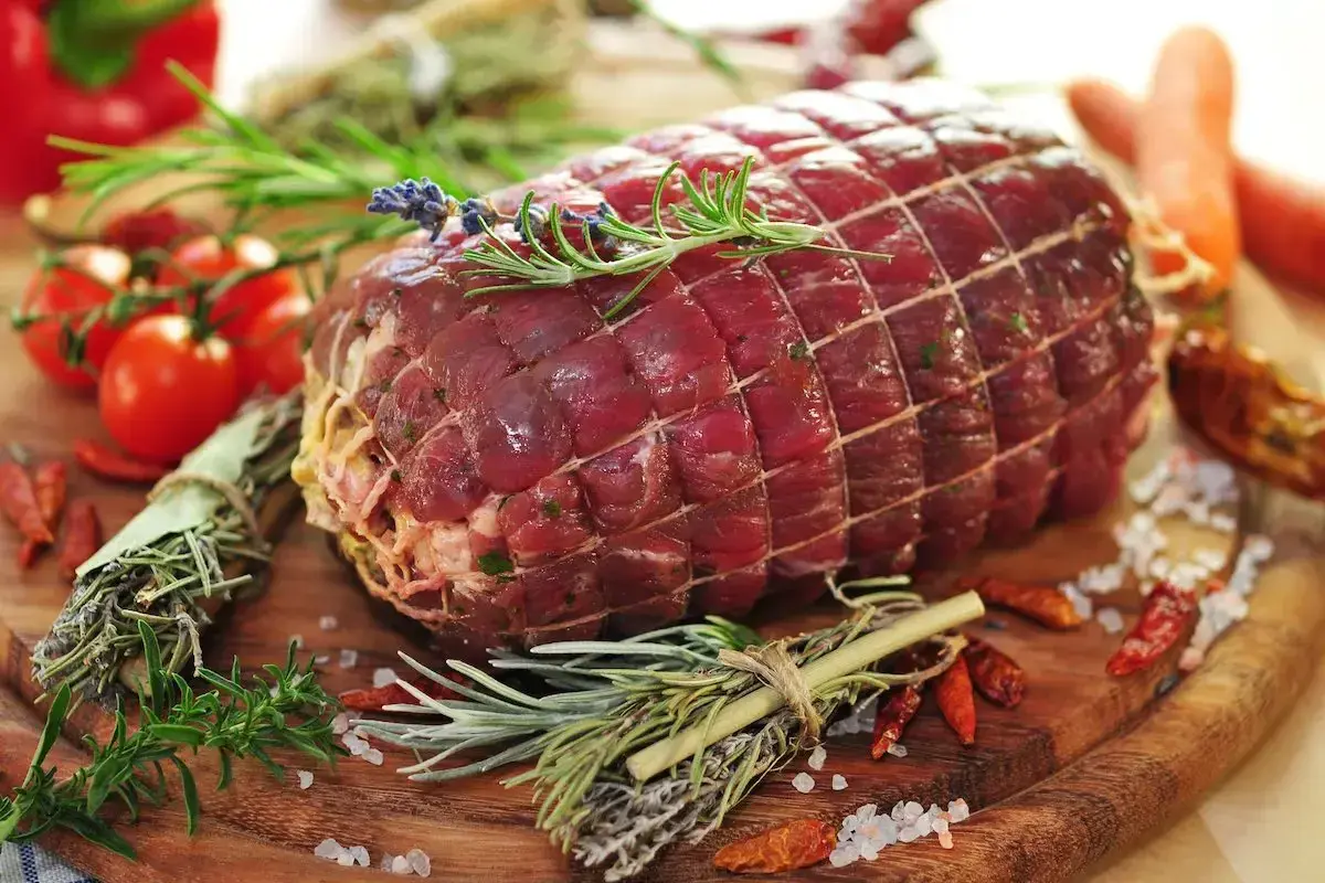 Un succulento arrosto di manzo, legato con spago e guarnito con rosmarino e lavanda, pronto per essere cucinato con pomodorini, peperoncini e carote.