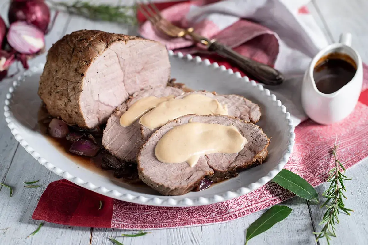 Arrosto di vitello ricetta della nonna, succulento e affettato, nappato con salsa cremosa. Servito su un piatto con cipolle rosse e contorno di salsa.