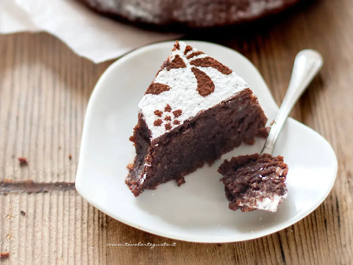 Una fetta di torta caprese ricetta facile, cosparsa di zucchero a velo e cacao, su un piatto bianco a forma di cuore.