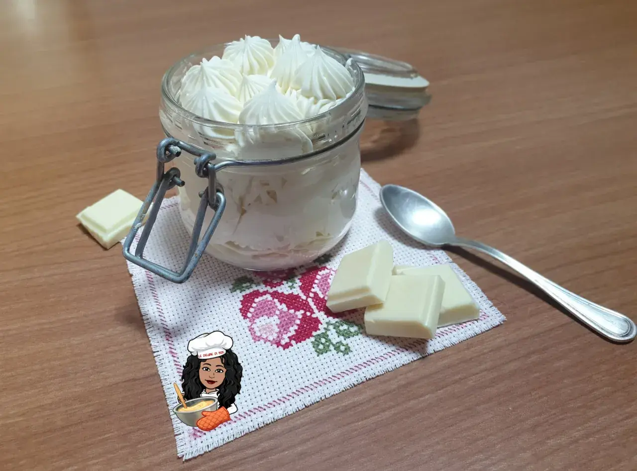 Un vasetto pieno di namelaka al cioccolato bianco, con pezzi di cioccolato e un cucchiaino.
