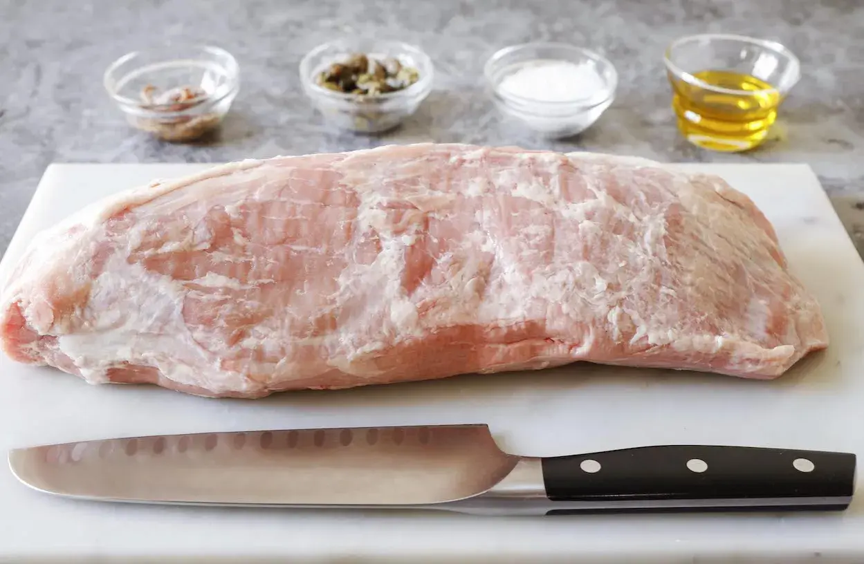 Un pezzo di magatello di vitello crudo, pronto per essere cucinato, con coltello e ciotoline di condimenti sullo sfondo.