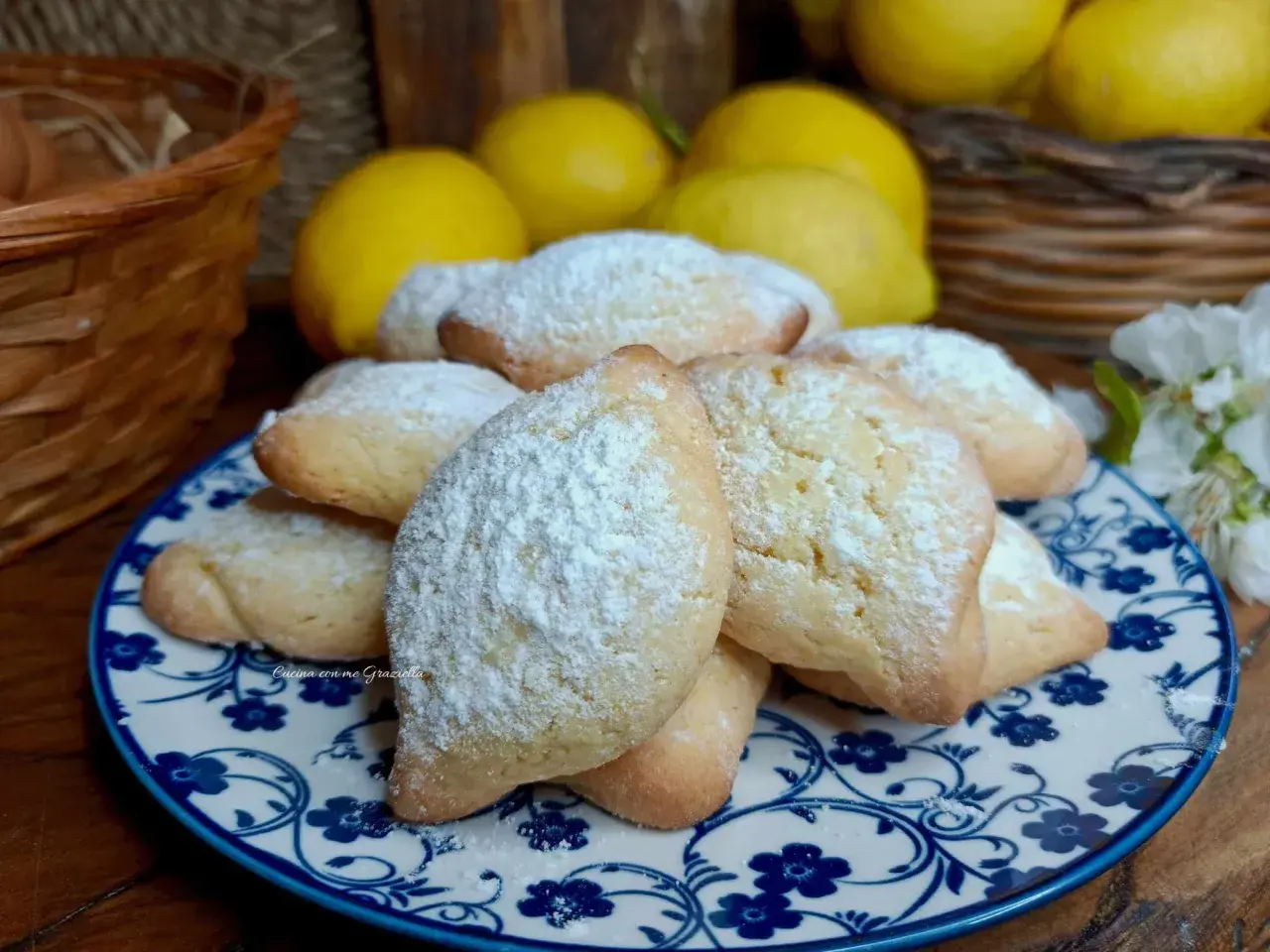 Biscotti con farina di mandorle, spolverati di zucchero a velo, su un piatto decorato. Sullo sfondo limoni e un cestino.