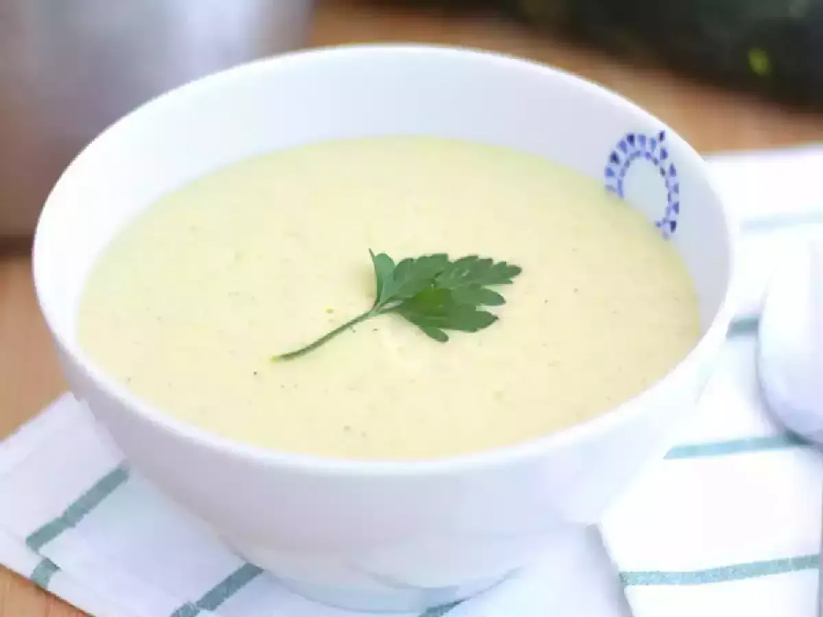 Una cremosa vellutata di zucchine, guarnita con un rametto di prezzemolo, servita in una ciotola bianca con un bordo decorato.