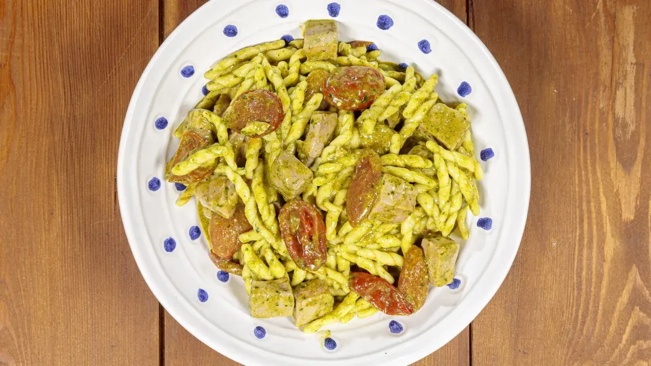 Piatto di trofie alla carlofortina con tonno, pomodorini secchi e pesto, servite su un piatto bianco con decorazioni blu.