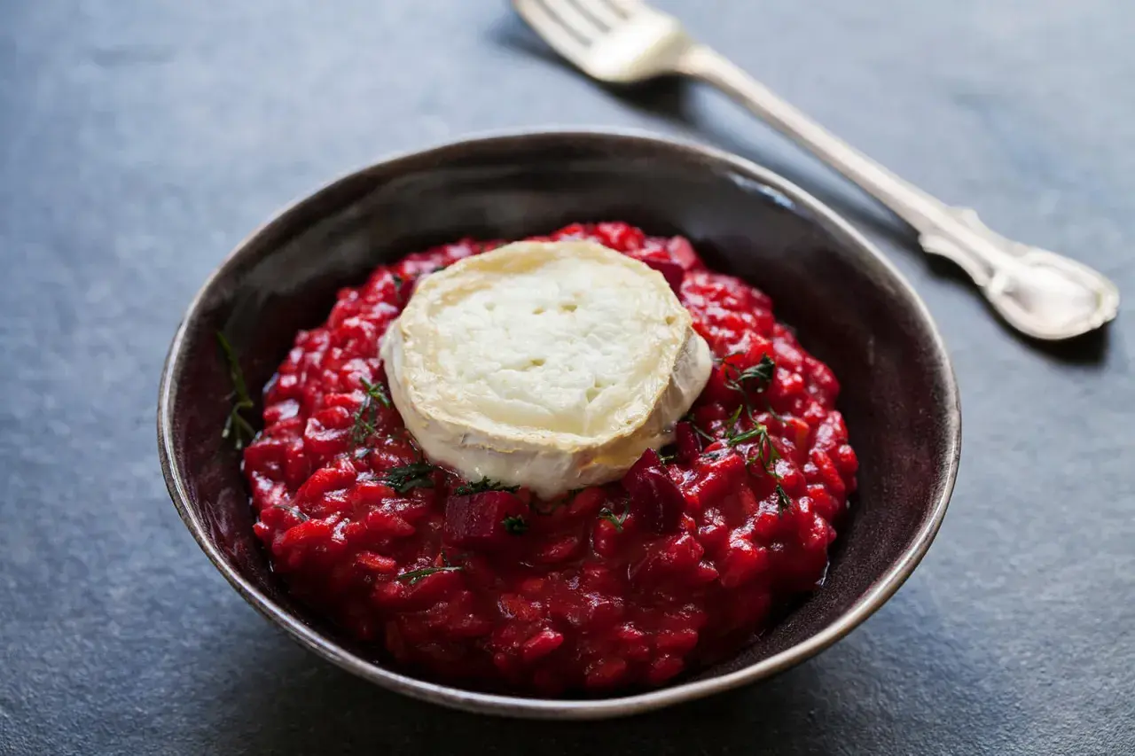 Un cremoso risotto alla barbabietola, con una fetta di formaggio di capra fuso sopra e un rametto di erbe aromatiche.
