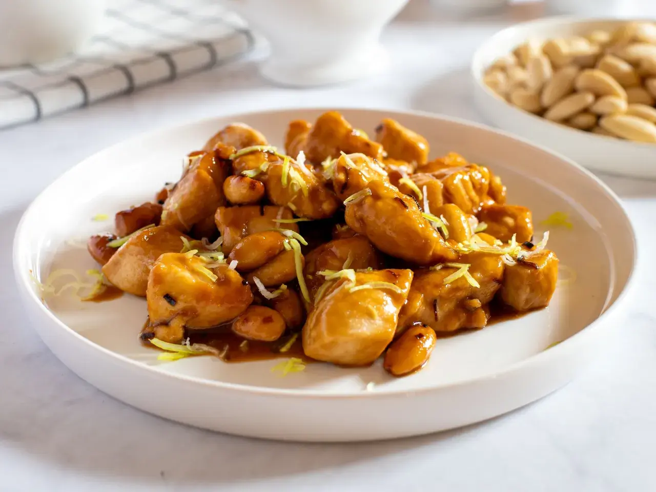Un invitante piatto di pollo alle mandorle, lucido e dorato, guarnito con erba cipollina. Sullo sfondo, una ciotola di mandorle intere.
