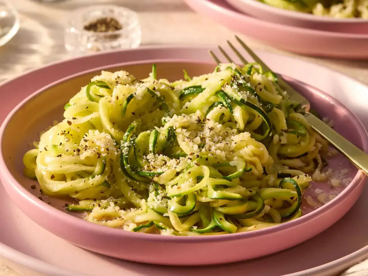 Un piatto di spaghetti di zucchine freschi, conditi con parmigiano e pepe nero, serviti su un piatto rosa.