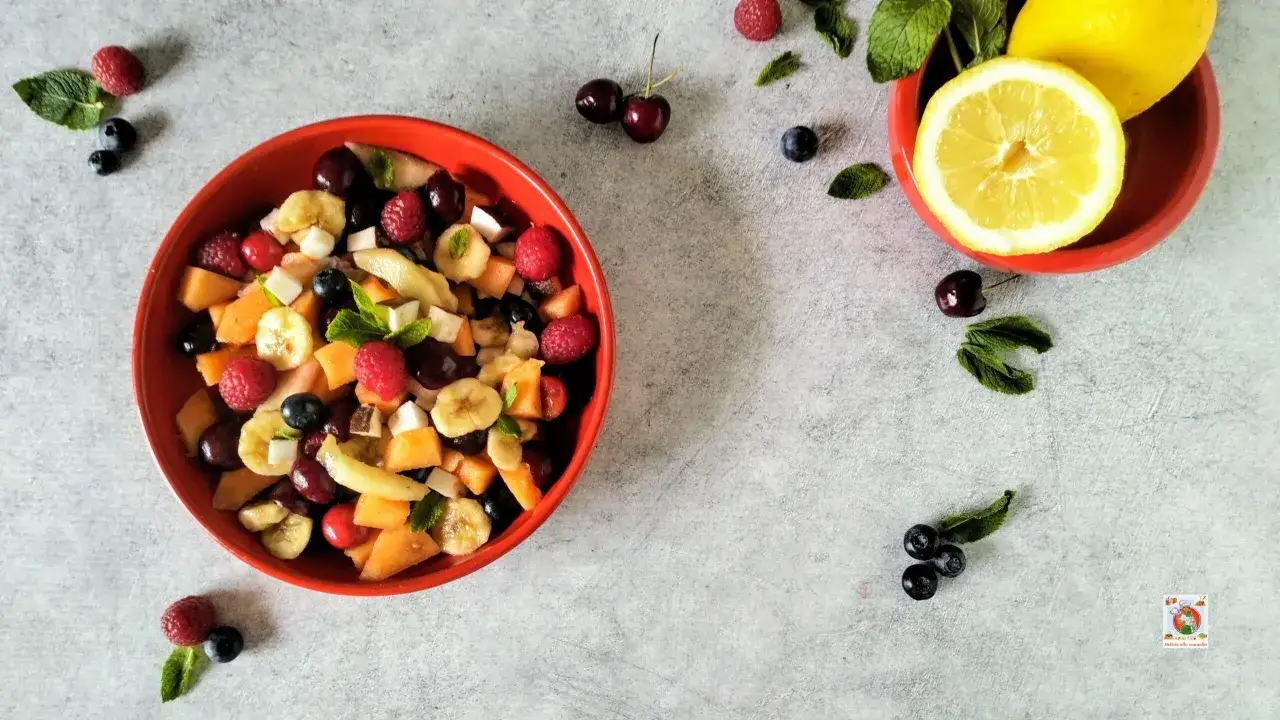 Una ciotola rossa piena di macedonia di frutta colorata, con lamponi, mirtilli, ciliegie, melone e banane. Accanto, limone e menta.