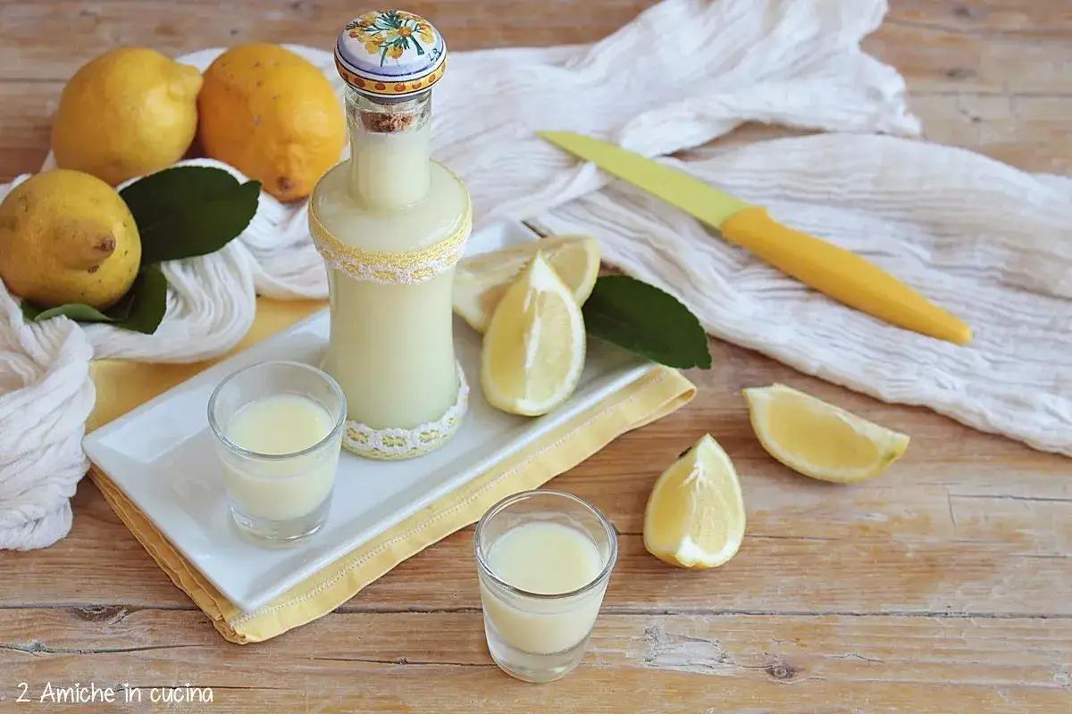 Una bottiglia e due bicchierini di cremosa crema di limoncello, circondati da limoni freschi e spicchi profumati.