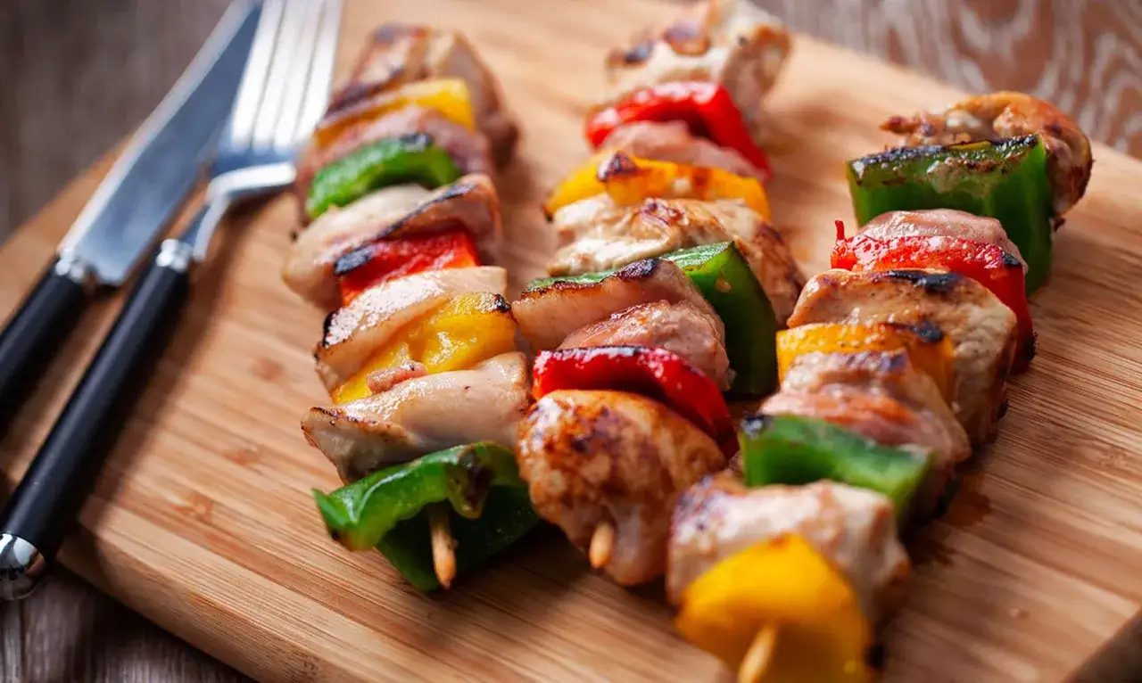 Spiedini di carne succulenti e colorati, alternati a peperoni verdi, rossi e gialli, pronti per essere gustati su un tagliere di legno.