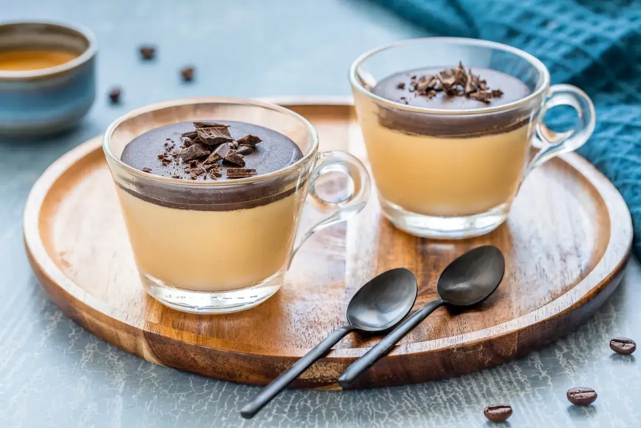 Due bicchierini di semifreddo al caffè, guarniti con cioccolato fondente e serviti su un vassoio di legno con cucchiaini.