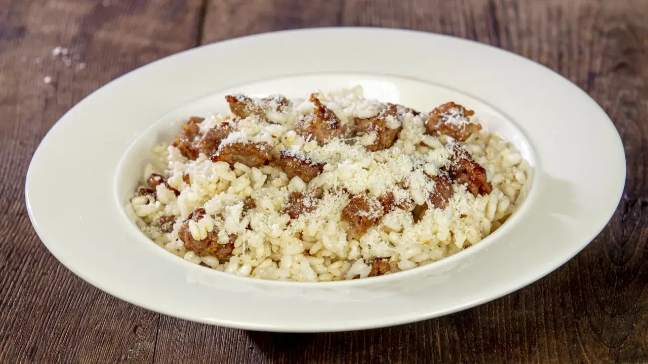 Un cremoso risotto alla pilota, cosparso di formaggio grattugiato e pezzetti di salsiccia, servito in un piatto bianco su un tavolo di legno scuro.