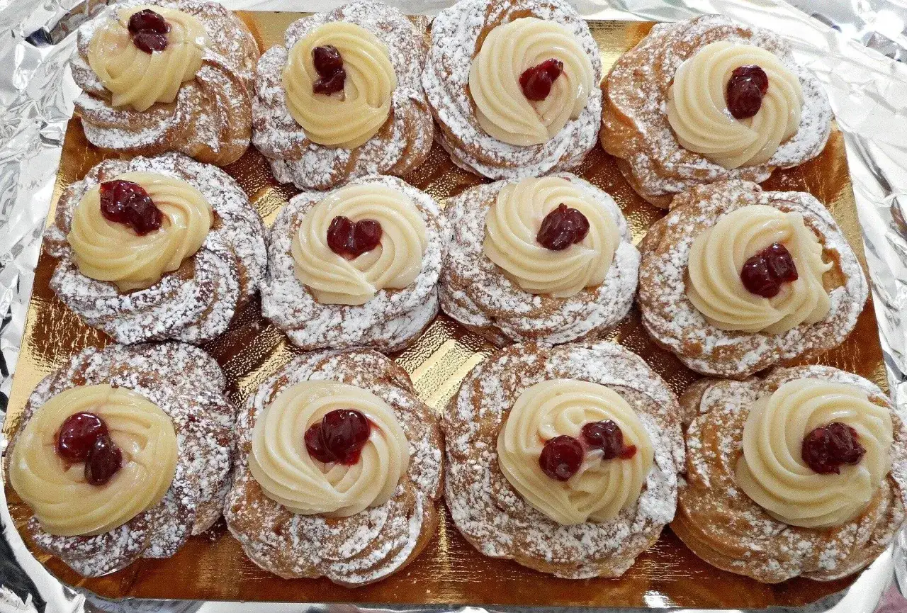 Un vassoio dorato pieno di zeppole di San Giuseppe, cosparse di zucchero a velo e decorate con crema pasticcera e ciliegie.