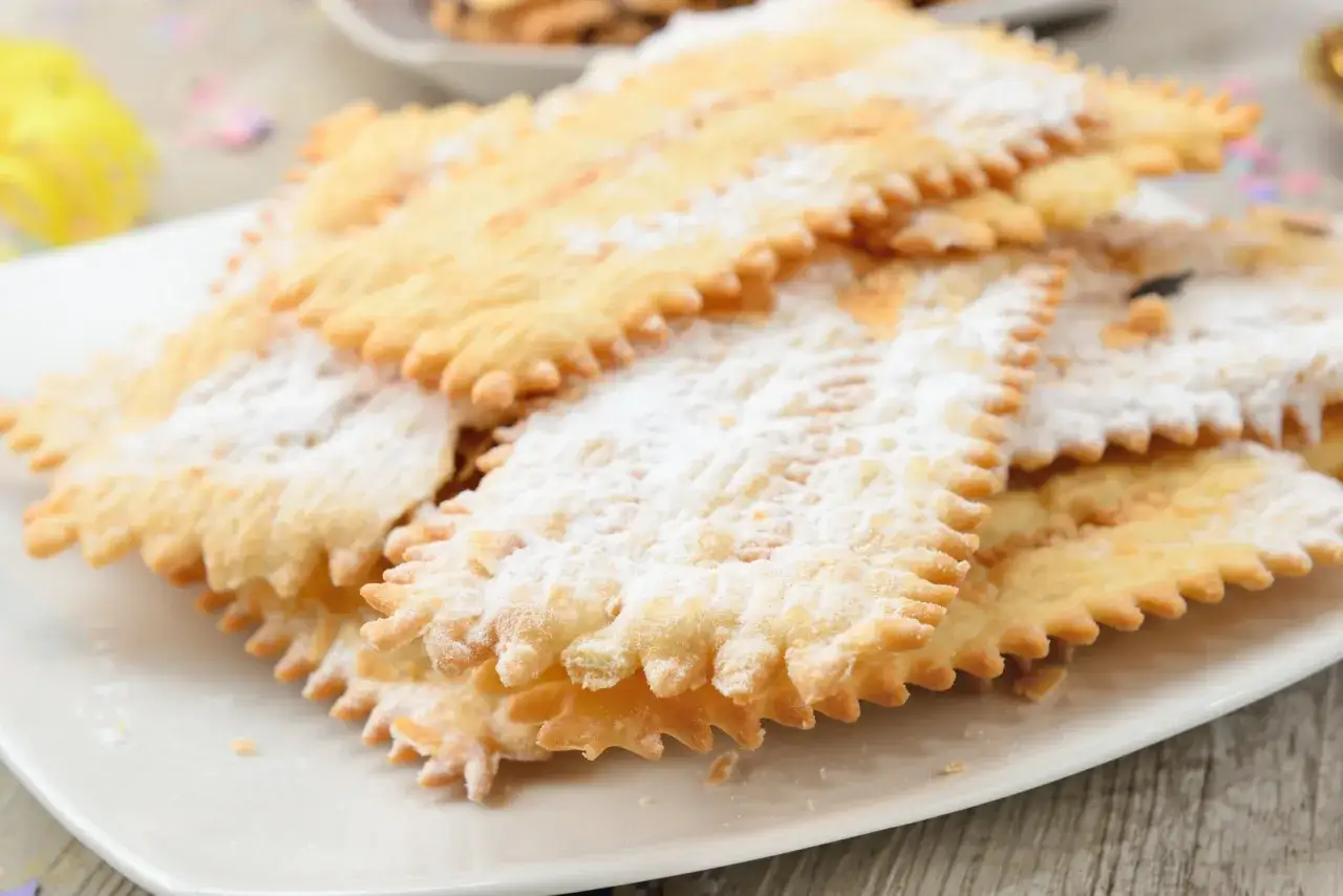 Un mucchio di chiacchiere, dolci senza glutine, cosparse di zucchero a velo su un piatto bianco.
