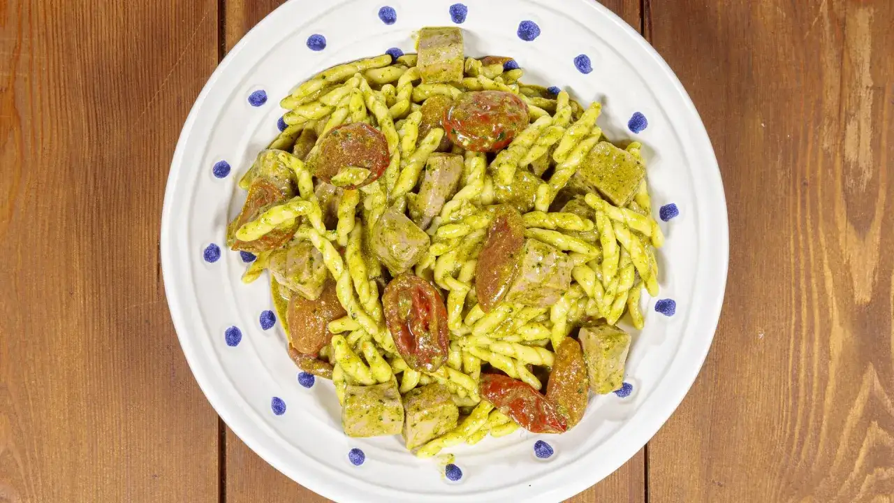 Piatto di trofie alla carlofortina con tonno, pomodorini secchi e pesto, servite su un piatto bianco con decorazioni blu.