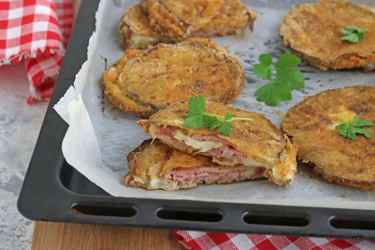 Cordon bleu di melanzane dorati e croccanti, ripieni di prosciutto e formaggio filante, guarniti con prezzemolo fresco su una teglia.