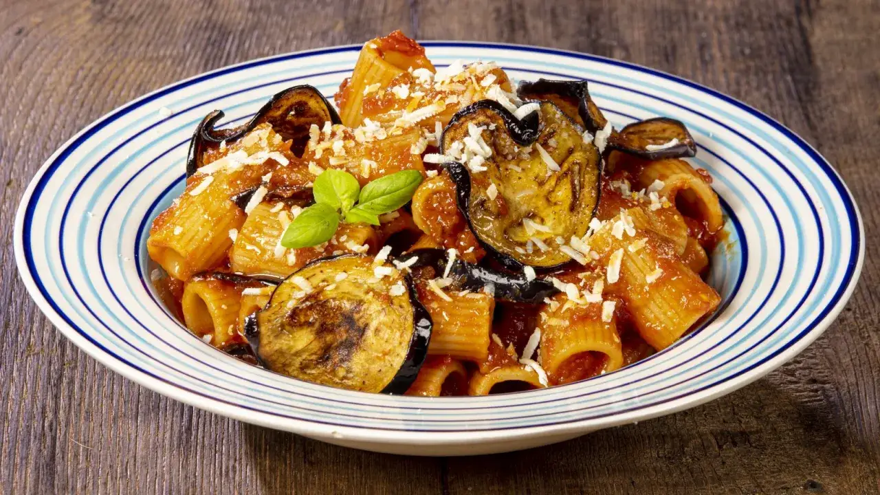 Un piatto di pasta alla norma fumante, con melanzane fritte, sugo rosso e ricotta salata grattugiata, guarnito con basilico fresco.