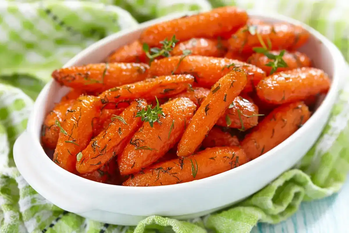 Carote in agrodolce glassate, guarnite con erbe aromatiche fresche, servite in una ciotola bianca su un tovagliolo verde.