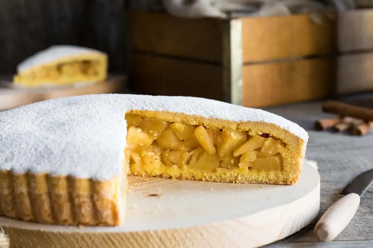 Una fetta di crostata di mele, cosparsa di zucchero a velo, rivela un ripieno succoso e dorato. Sullo sfondo, bastoncini di cannella e una cassetta di legno.