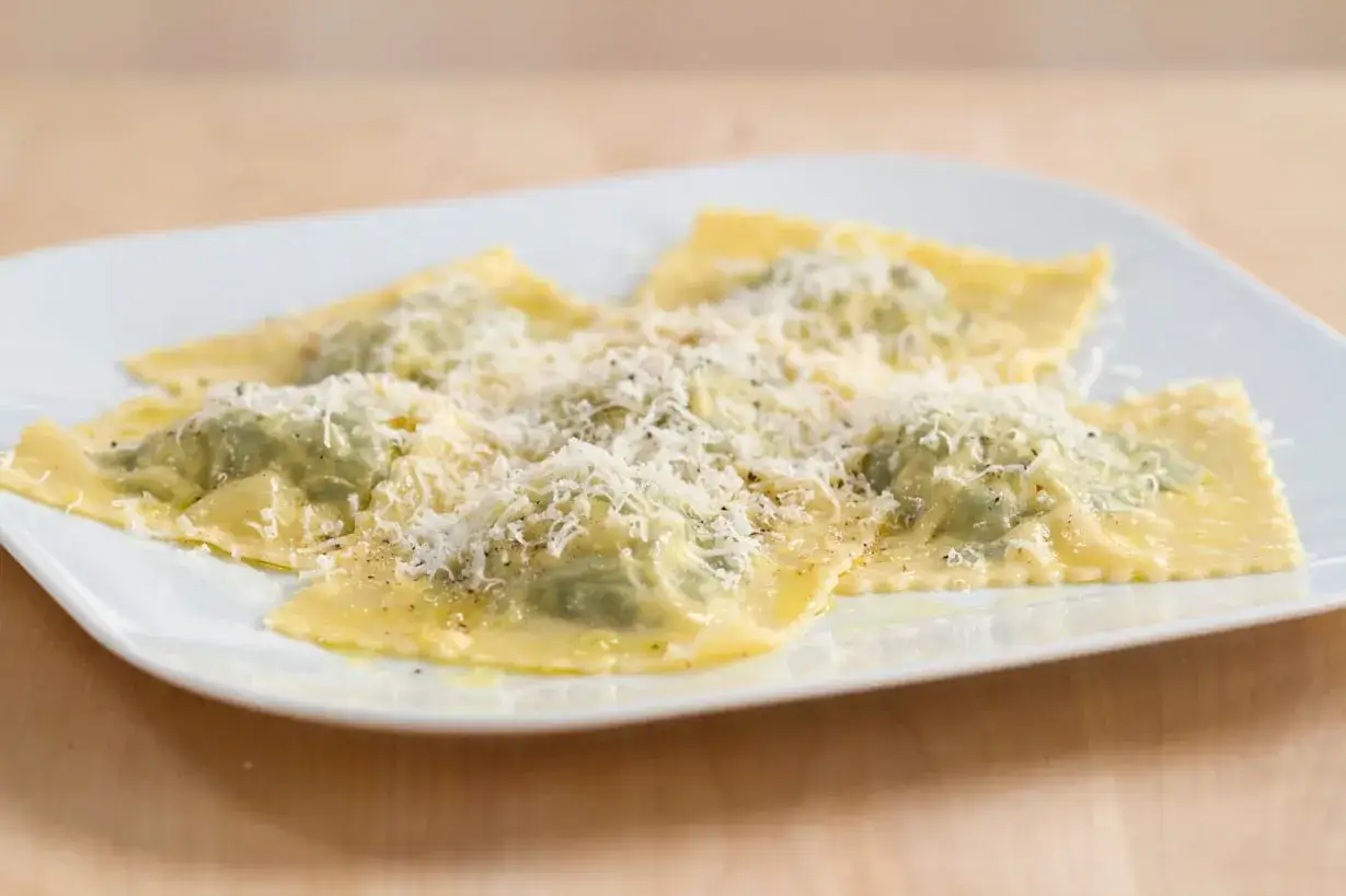 Ravioli ricotta e spinaci su piatto bianco, conditi con parmigiano grattugiato e pepe.