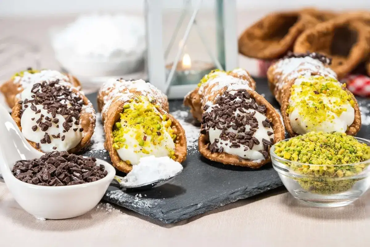 Cannoli siciliani ricetta: deliziosi dolci con crema e granella di pistacchio o gocce di cioccolato, spolverati di zucchero a velo.