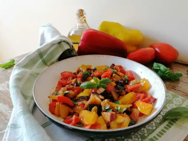 Un piatto colorato di peperonata con melanzane, pomodori e peperoni, guarnito con basilico fresco. La ricetta perfetta per un contorno estivo.