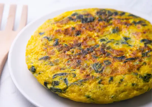 Frittata di spinaci dorata e soffice su un piatto bianco, con un utensile in legno sullo sfondo.