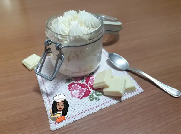 Un vasetto pieno di namelaka al cioccolato bianco, con pezzi di cioccolato e un cucchiaino.