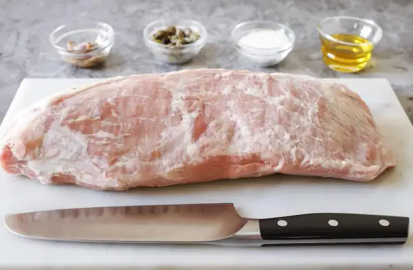 Un pezzo di magatello di vitello crudo, pronto per essere cucinato, con coltello e ciotoline di condimenti sullo sfondo.
