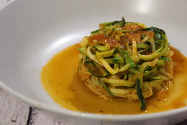 Un piatto di spaghetti di zucchine conditi con sugo rosso, serviti in una ciotola bianca.