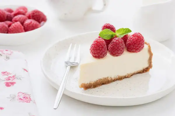 Una fetta di torta fredda allo yogurt, guarnita con lamponi freschi e menta, su un piatto bianco.