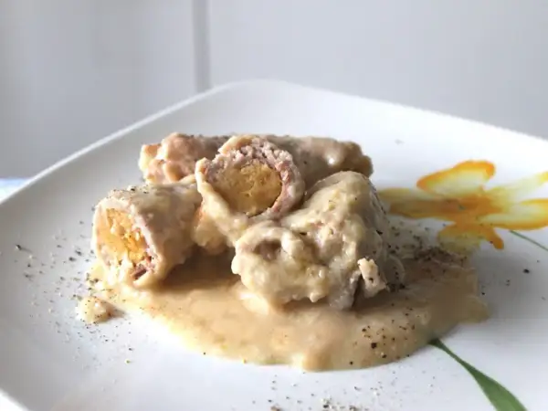 Involtini di carne succulenti in salsa cremosa, cosparsi di pepe nero su un piatto bianco decorato con un fiore giallo.