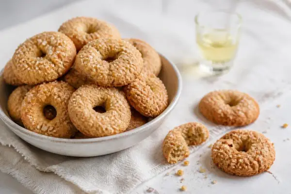 Ciambelline al vino friabilissime, dorate e cosparse di zucchero, in una ciotola e sparse su un panno, con un bicchierino di vino.