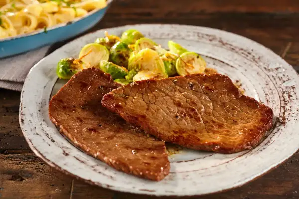 Due fette di paillard di vitello succulento servite con cavolini di Bruxelles arrostiti e un contorno di pasta in un piatto rustico.