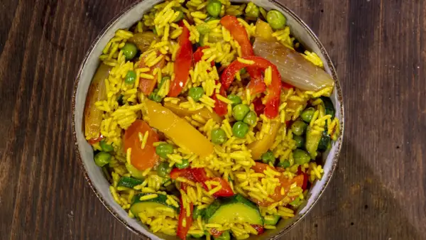 Un invitante riso al curry con piselli, peperoni rossi e gialli, zucchine e cipolla, servito in una ciotola su un tavolo di legno.
