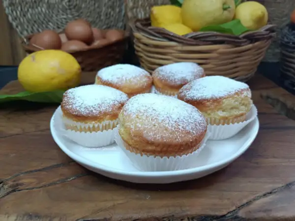 Cinque muffin allo yogurt spolverati di zucchero a velo, pronti per essere gustati. Sullo sfondo, limoni e uova fresche.