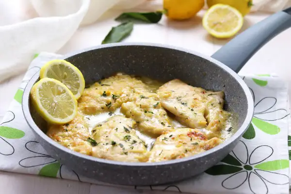 Scaloppine al limone cremose in padella, guarnite con spicchi di limone fresco e prezzemolo tritato.