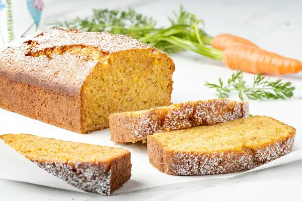 Torta carote e mandorle soffice e dorata, cosparsa di zucchero a velo. Due fette sono servite accanto al pane intero, con carote fresche sullo sfondo.